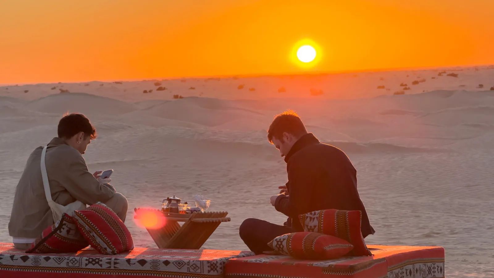 Two travellers sharing tea on a rug at sunset