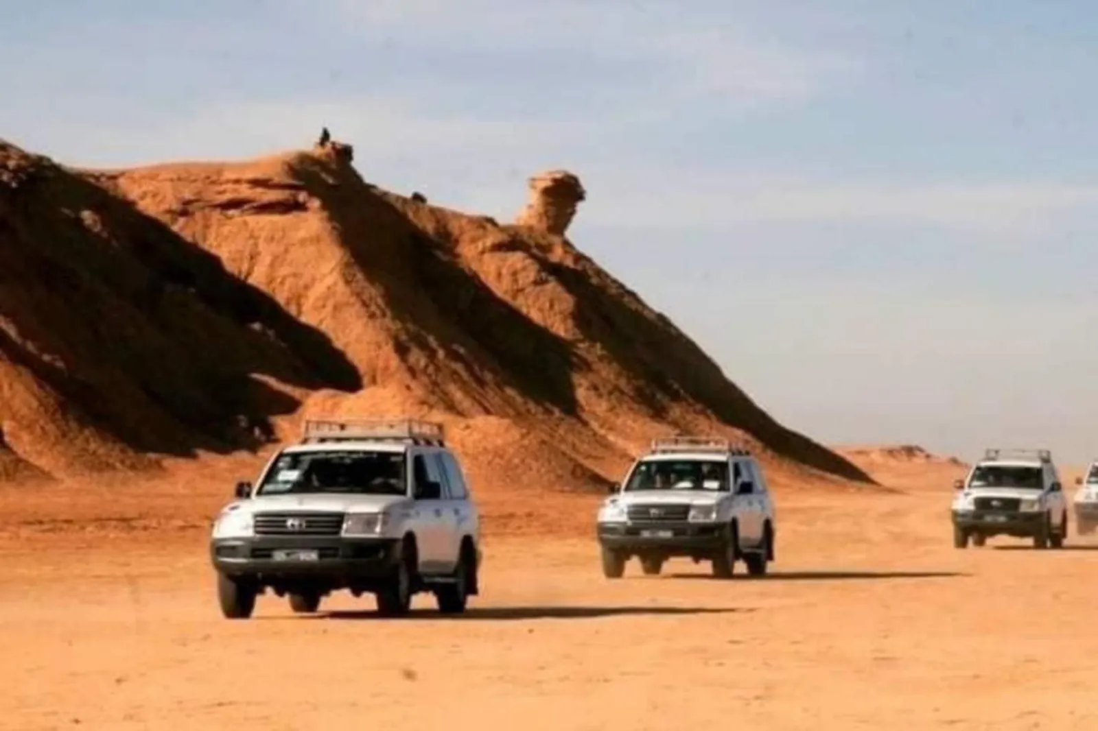 A convoy of 4x4s crossing open desert past a sandstone outcrop